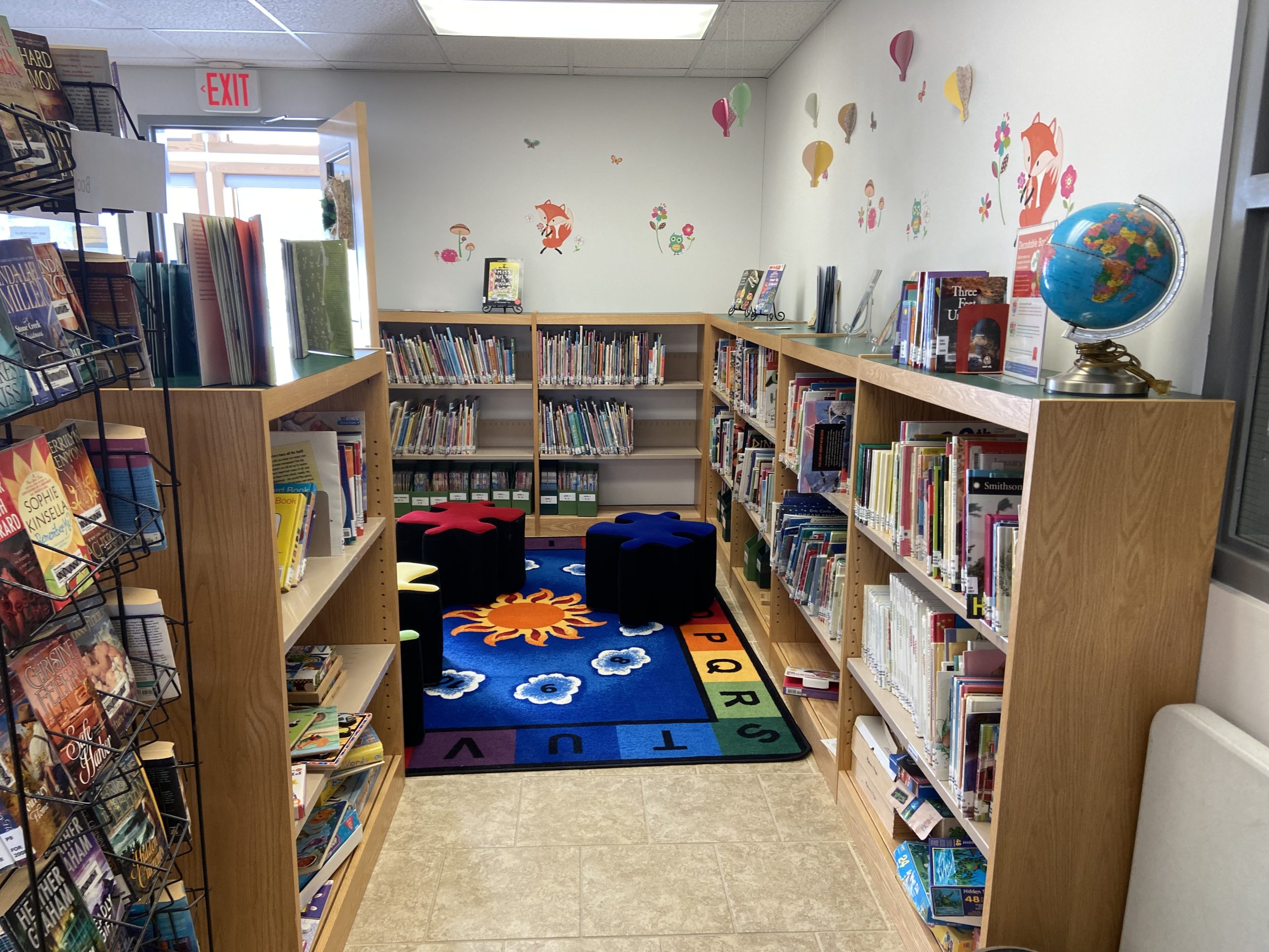 A view of the kid's corner within the library. It is a small area, walled off by bookshelves filled with various children's books, with a carpet in the middle and puzzle-piece shaped chairs. There are decorations placed on the walls and atop the bookshelves.