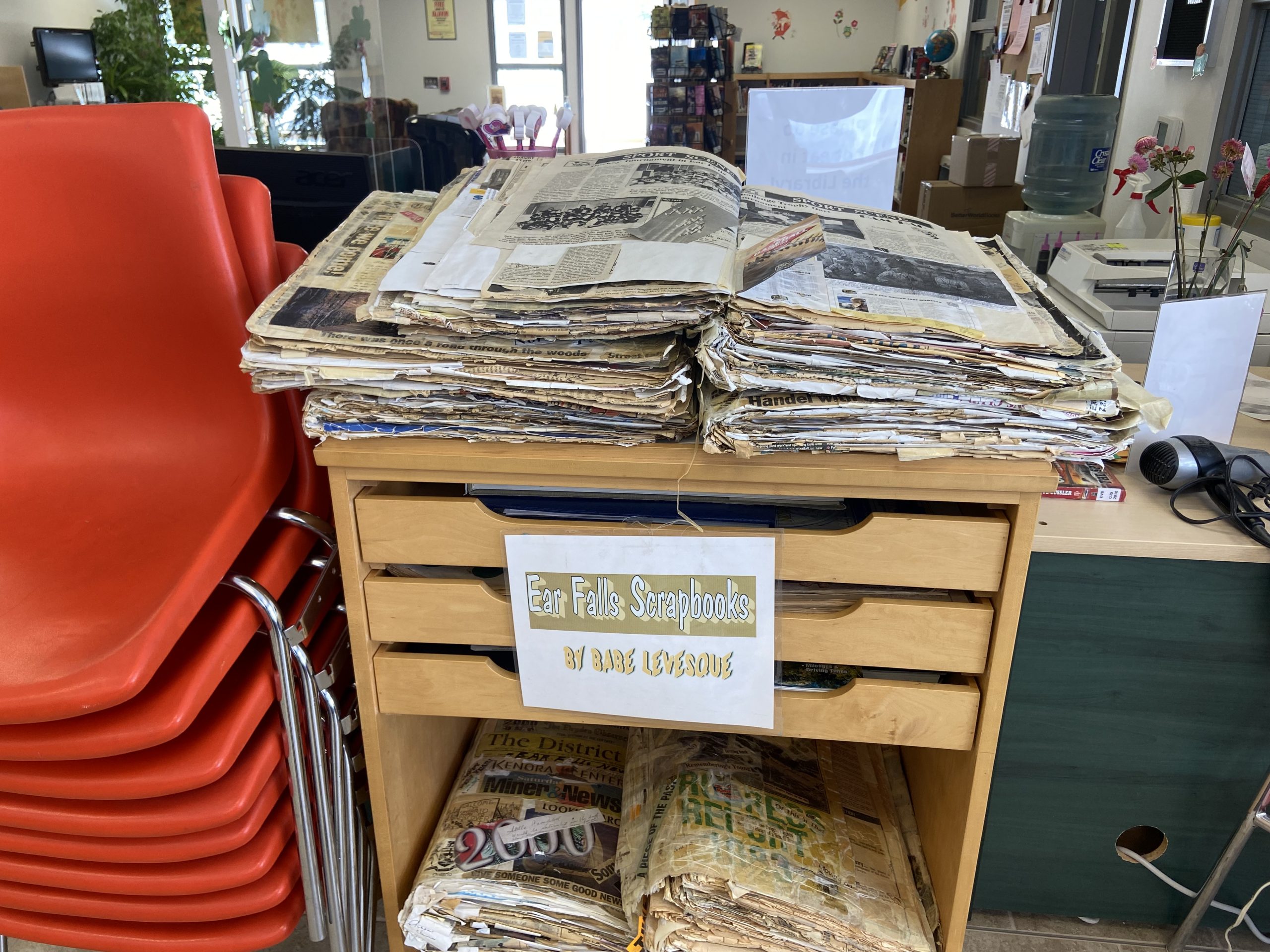 Various old newspapers and various other paper to be recycled lines a desk. A sign is placed on the front reading "Ear Falls Scrapbooks by Babe Levesque".
